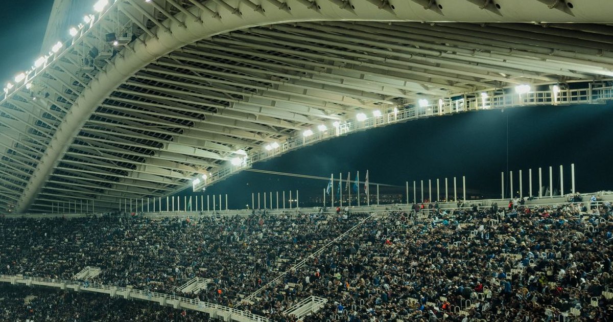 Packed stadium stands at night under a curved white roof lit by rows of floodlights, with thousands of fans filling the seats