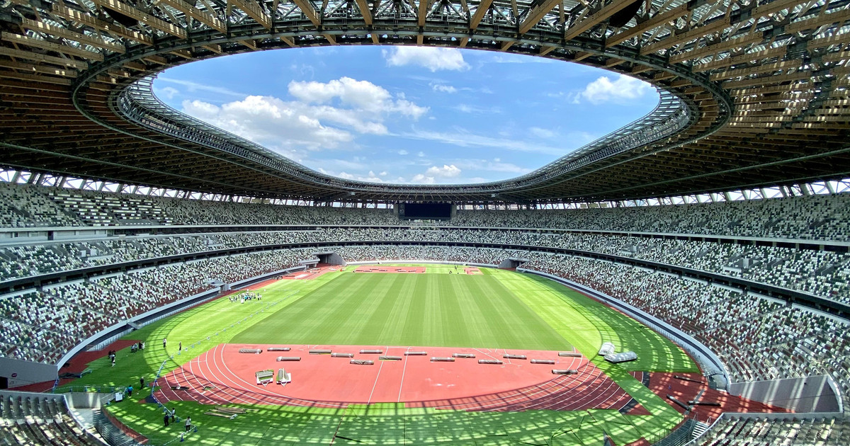 Wide view inside an empty stadium on a sunny day, with rows of seats circling a green field and red running track under an open oval roof while a few crew members work on the track