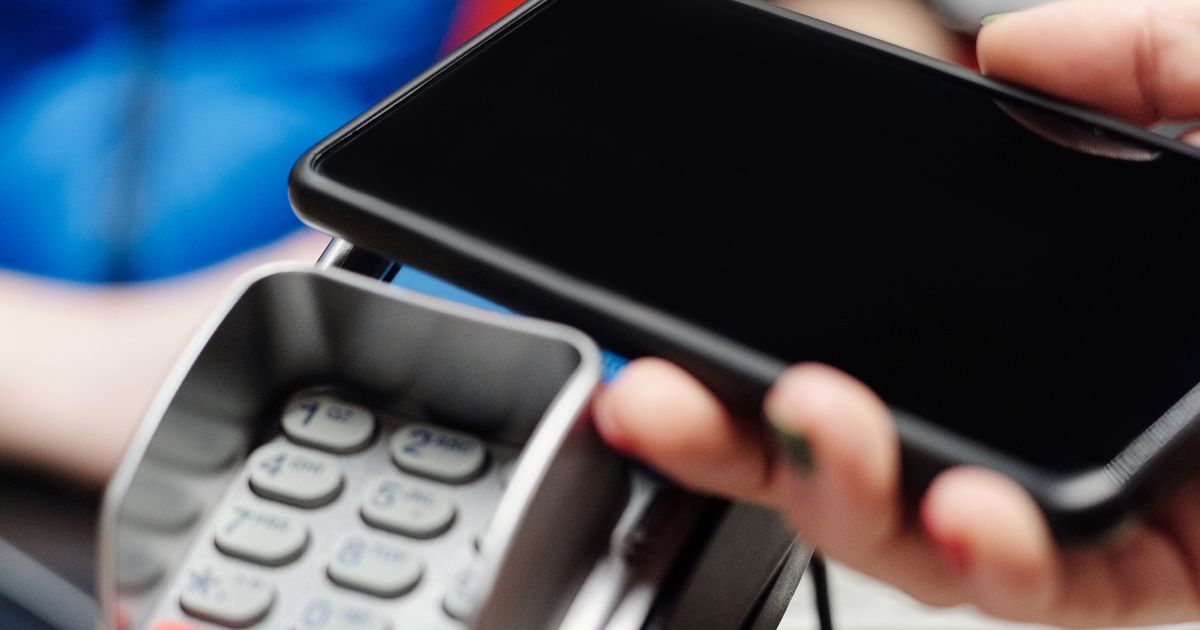 Close view of a hand holding a smartphone over a card reader keypad to make a contactless payment, with a blurred blue shirt in the background