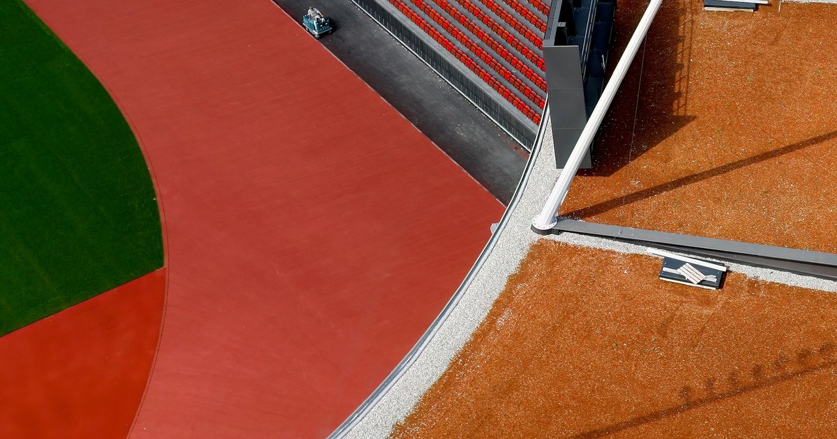Aerial view of an empty athletics stadium with a curved red running track, green infield, rows of red seats and tall floodlight poles casting long shadows